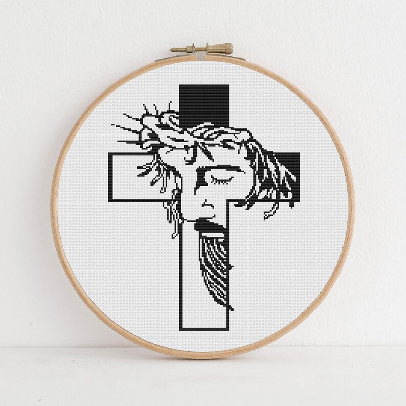 Puede incluir: Un bastidor de bordado circular de madera que contiene un patr&oacute;n de punto de cruz de una ilustraci&oacute;n gr&aacute;fica en blanco y negro. El dise&ntilde;o presenta un rostro estilizado con una corona de espinas sobre una cruz negra en fondo blanco.