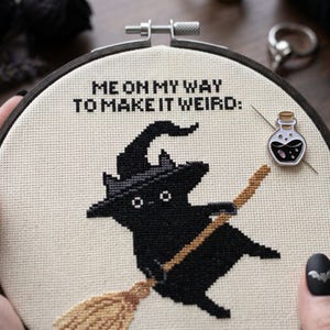 Puede incluir: Un proyecto de punto de cruz en un bastidor de madera circular que muestra un gato negro con sombrero de bruja sobre una escoba. El texto "ME ON MY WAY TO MAKE IT WEIRD:" est&aacute; bordado en negro. Un peque&ntilde;o pin de esmalte con forma de frasco est&aacute; sujeto.