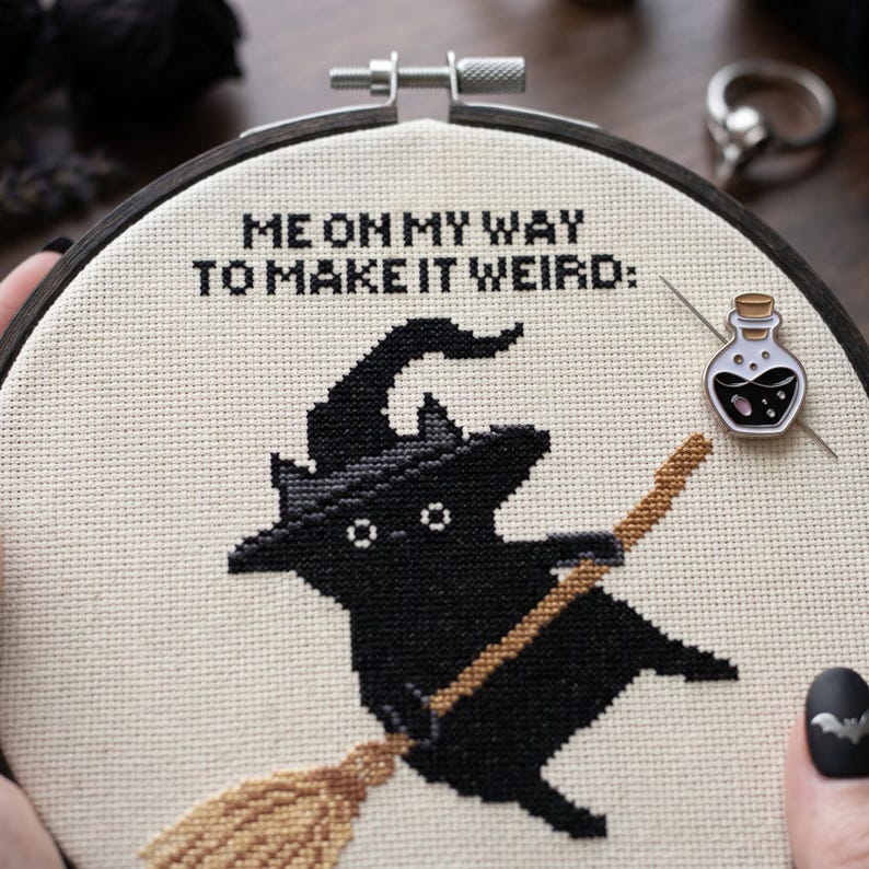 Puede incluir: Un proyecto de punto de cruz en un bastidor de madera circular que muestra un gato negro con sombrero de bruja sobre una escoba. El texto "ME ON MY WAY TO MAKE IT WEIRD:" est&aacute; bordado en negro. Un peque&ntilde;o pin de esmalte con forma de frasco est&aacute; sujeto.