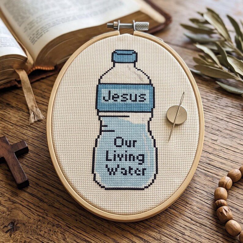 Puede incluir: Un bastidor de bordado ovalado de madera que contiene un patr&oacute;n de punto de cruz de una botella de agua. La botella est&aacute; bordada con hilo azul claro y negro con el texto Jesus Our Living Water. Un im&aacute;n para agujas est&aacute; sujeto a la tela.