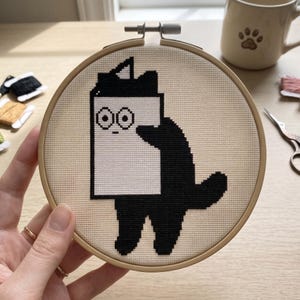 May include: A circular wooden embroidery hoop containing a cross-stitch design on cream-colored fabric. The design features a black cat silhouette holding a large white rectangular object with a simple black outline and two circular eyes.