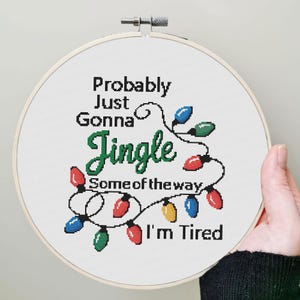 Może przedstawiać: Obręcz do haftu krzyżykowego z napisem "Probably Just Gonna Jingle Some of the way I'm Tired". Projekt zawiera kolorowe lampki choinkowe w kolorze czerwonym, zielonym, niebieskim i ż&oacute;łtym. Obręcz jest trzymana przez osobę.