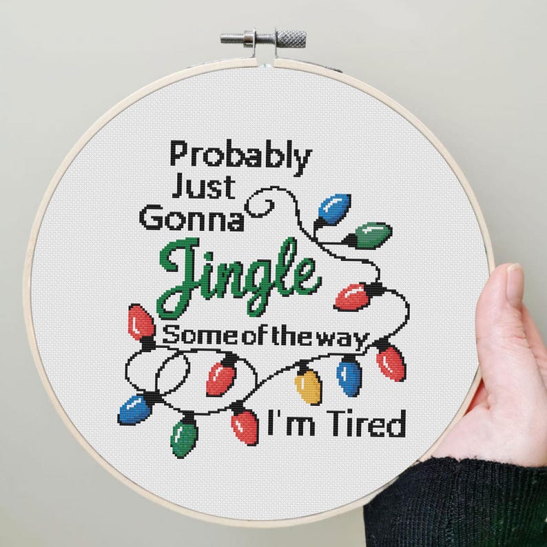 Może przedstawiać: Obręcz do haftu krzyżykowego z napisem "Probably Just Gonna Jingle Some of the way I'm Tired". Projekt zawiera kolorowe lampki choinkowe w kolorze czerwonym, zielonym, niebieskim i ż&oacute;łtym. Obręcz jest trzymana przez osobę.