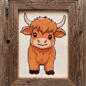 May include: Cross-stitch artwork of a Highland cow in a rustic wooden frame. The cow is depicted in shades of orange and brown, with a smiling face and curved horns. The frame is made of reclaimed wood with metal corner brackets.