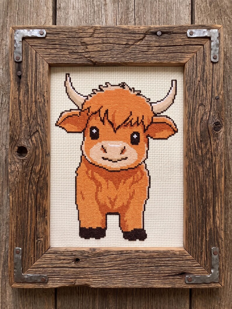 May include: Cross-stitch artwork of a Highland cow in a rustic wooden frame. The cow is depicted in shades of orange and brown, with a smiling face and curved horns. The frame is made of reclaimed wood with metal corner brackets.