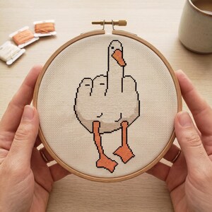 Peut inclure: Un projet de broderie au point de croix dans un tambour en bois circulaire repr&eacute;sentant une oie blanche au bec et aux pattes orange, montrant son aile lev&eacute;e dans un geste obsc&egrave;ne. Le motif est brod&eacute; sur un tissu blanc &agrave; texture quadrill&eacute;e.