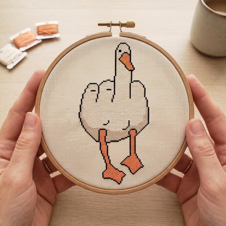 Peut inclure: Un projet de broderie au point de croix dans un tambour en bois circulaire repr&eacute;sentant une oie blanche au bec et aux pattes orange, montrant son aile lev&eacute;e dans un geste obsc&egrave;ne. Le motif est brod&eacute; sur un tissu blanc &agrave; texture quadrill&eacute;e.
