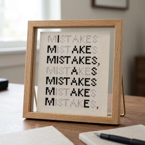 K&ouml;nnte beinhalten: Ein quadratischer Bilderrahmen aus hellem Holz mit einem wei&szlig;en Aida-Stoff, auf dem das Wort MISTAKES siebenmal in abwechselnd schwarzen und grauen pixeligen Buchstaben im Kreuzstich gestickt ist, inklusive zwei kleiner roter Kommas am Zeilenende.
