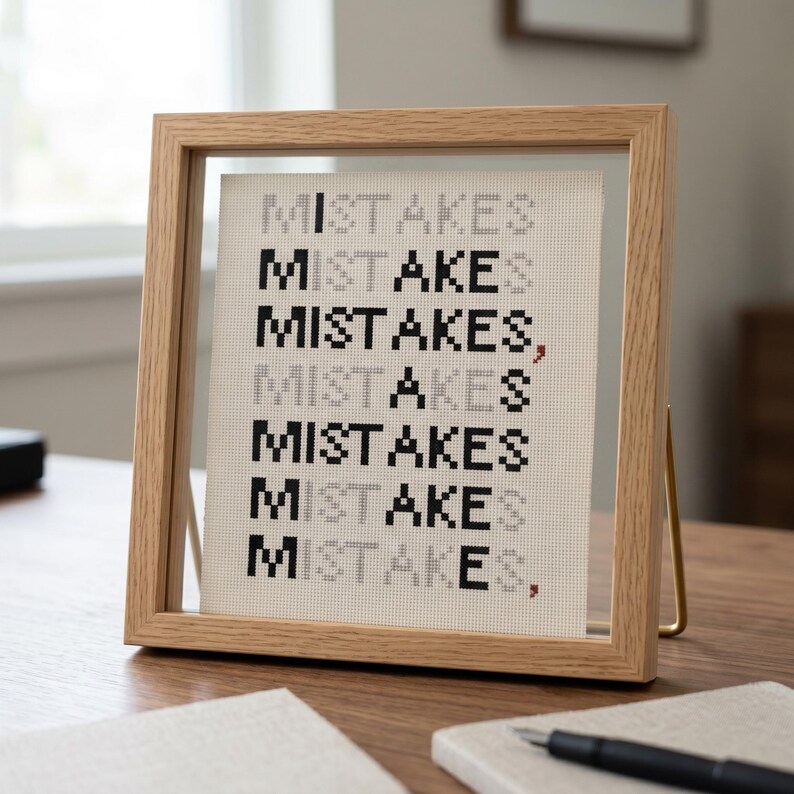 K&ouml;nnte beinhalten: Ein quadratischer Bilderrahmen aus hellem Holz mit einem wei&szlig;en Aida-Stoff, auf dem das Wort MISTAKES siebenmal in abwechselnd schwarzen und grauen pixeligen Buchstaben im Kreuzstich gestickt ist, inklusive zwei kleiner roter Kommas am Zeilenende.