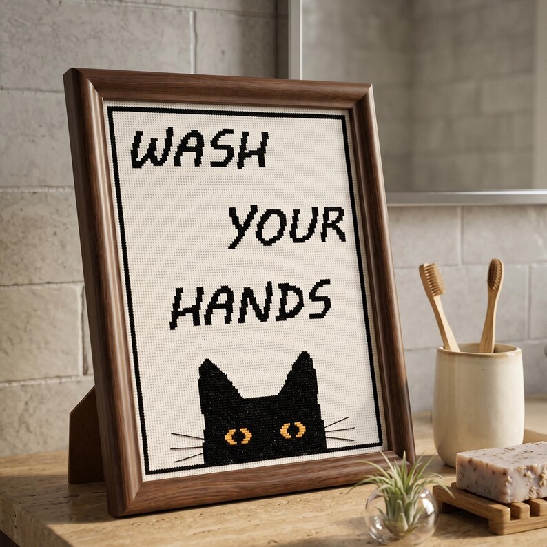 K&ouml;nnte beinhalten: Ein gerahmtes Kreuzstichbild mit dem schwarzen Text WASH YOUR HANDS &uuml;ber einem schwarzen Katzengesicht mit gelben Augen. Das Kunstwerk befindet sich in einem dunkelbraunen Holzrahmen und zeigt ein klares, pixeliges Design auf hellem Stoff.