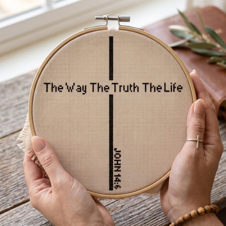 May include: A circular wooden embroidery hoop containing beige fabric with black cross-stitch embroidery. The design features a vertical black line forming a cross with the text The Way The Truth The Life and the reference JOHN 14:6.