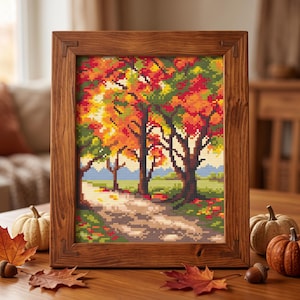 Può includere: Un ricamo a punto croce che raffigura un paesaggio autunnale con alberi dai colori vivaci rosso, arancione e giallo lungo un sentiero, incorniciato in una cornice di legno rettangolare. La scena presenta un cielo azzurro e erba verde.