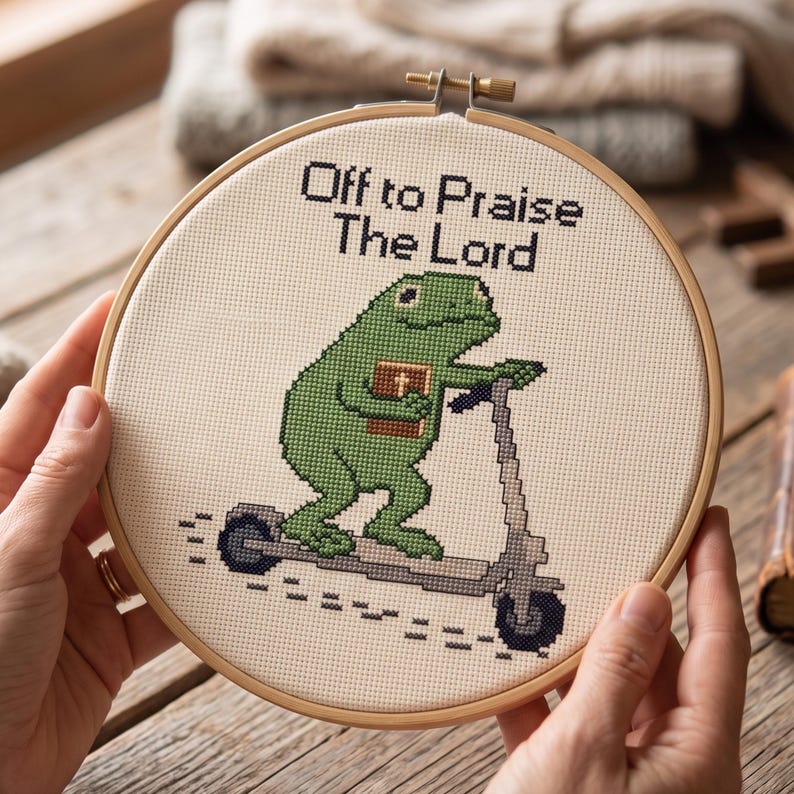 May include: A circular wooden embroidery hoop containing a cross-stitch design on cream fabric. The design features a green frog riding a grey scooter while holding a brown book with a cross on the cover. The text reads Off to Praise The Lord.