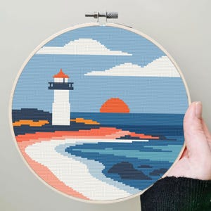 May include: A cross-stitch embroidery hoop featuring a coastal scene. The design includes a white lighthouse with a red roof, a setting sun, ocean waves, and clouds. The color palette includes shades of blue, orange, and white.