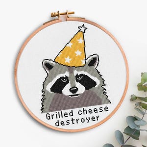 Op de afbeelding: Kruissteekborduurwerk van een wasbeer met een gele feesthoed met witte sterren, in een houten borduurring. De tekst "Grilled cheese destroyer" staat onder de wasbeer.