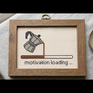 Puede incluir: Un bordado de punto de cruz en un marco de madera rectangular que muestra una cafetera italiana gris vertiendo caf&eacute; marr&oacute;n en una barra de carga. Debajo de la barra, el texto dice motivation loading... en letras min&uacute;sculas negras.