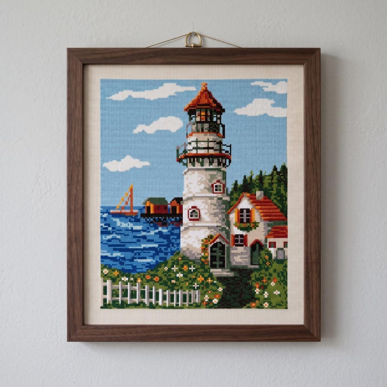 Peut inclure: Une broderie au point de croix encadr&eacute;e repr&eacute;sentant une sc&egrave;ne c&ocirc;ti&egrave;re avec un phare blanc, une petite maison au toit rouge, une mer bleue avec un voilier et une cl&ocirc;ture blanche. L'&oelig;uvre est dans un cadre en bois brun fonc&eacute; avec une attache.