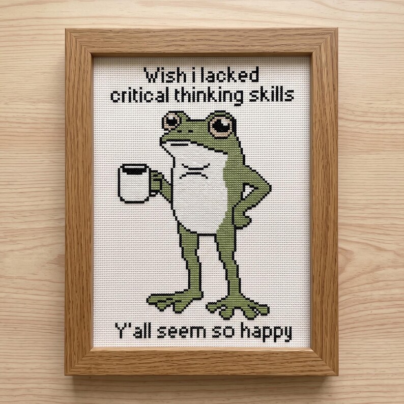 May include: A framed cross-stitch embroidery featuring a green frog holding a white mug. The text above reads Wish I lacked critical thinking skills and the text below reads Y'all seem so happy. The artwork is enclosed in a light brown wooden frame.
