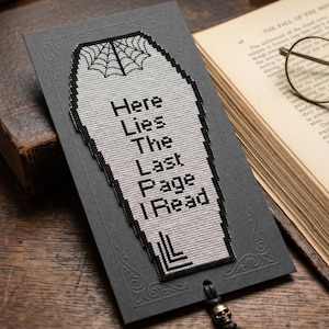 Pode incluir: Um marcador de página cinza com um design de caixão e uma teia de aranha. O texto "Here Lies The Last Page I Read" é exibido. Um pingente de caveira pende da parte inferior. O marcador de página está ao lado de um livro aberto.