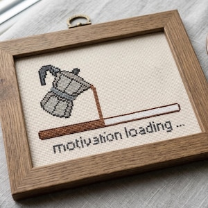 Puede incluir: Un bordado de punto de cruz en un marco de madera que muestra una cafetera italiana gris vertiendo caf&eacute; marr&oacute;n en una barra de carga horizontal. Debajo de la barra, el texto dice motivation loading... en letras min&uacute;sculas negras sobre tela crema.
