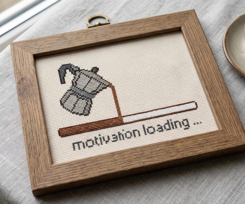 Puede incluir: Un bordado de punto de cruz en un marco de madera que muestra una cafetera italiana gris vertiendo caf&eacute; marr&oacute;n en una barra de carga horizontal. Debajo de la barra, el texto dice motivation loading... en letras min&uacute;sculas negras sobre tela crema.