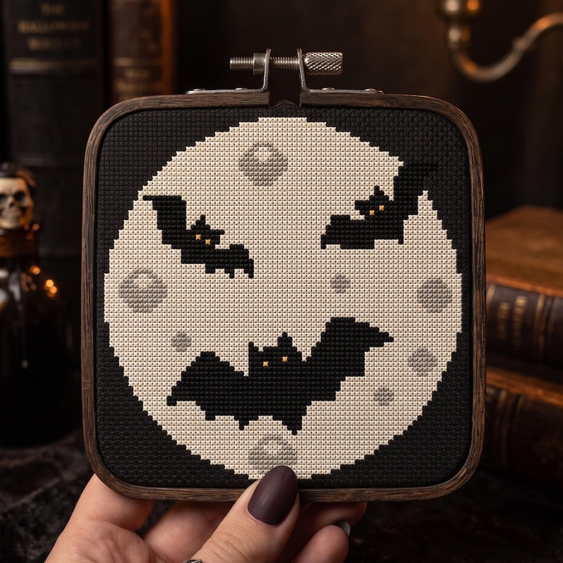 Pu&ograve; includere: Un telaio da ricamo quadrato in legno contenente un disegno a punto croce di una grande luna circolare pallida su sfondo nero. Tre pipistrelli neri con piccoli occhi gialli sono posizionati sulla luna, che presenta sottili crateri circolari grigi.
