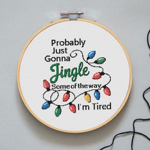 Może przedstawiać: Obręcz do haftu krzyżykowego z napisem "Probably Just Gonna Jingle Some of the way I'm Tired". Projekt zawiera kolorowe lampki choinkowe w kolorze czerwonym, zielonym, niebieskim i ż&oacute;łtym. Obręcz jest na jasnoszarym tle.