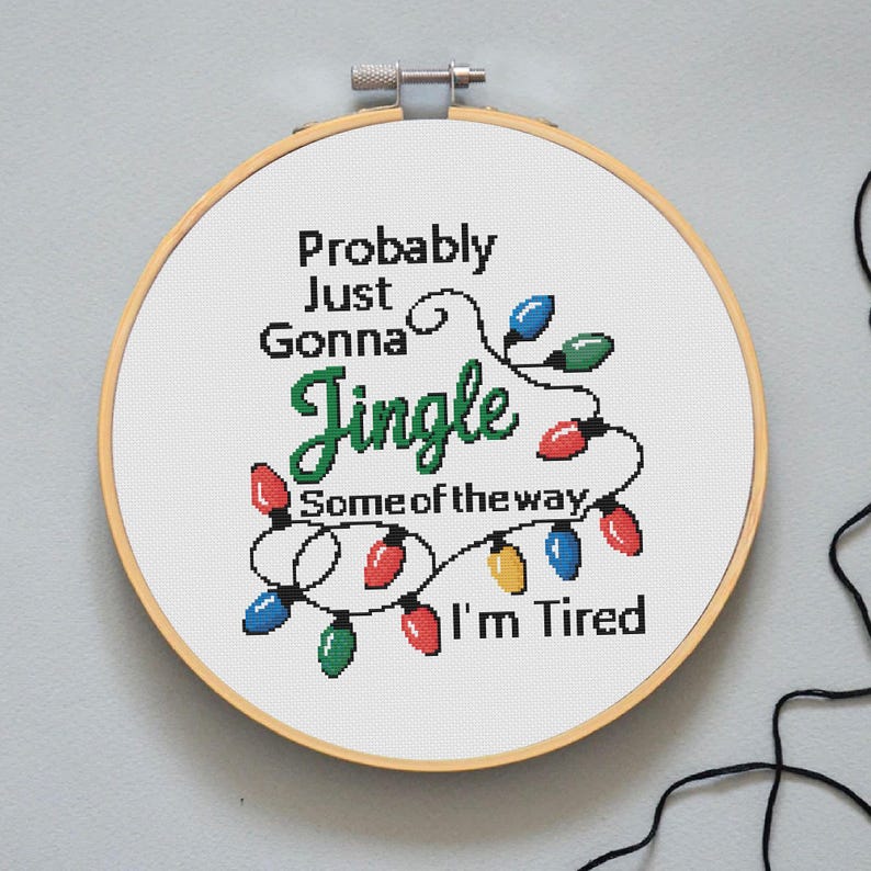 Może przedstawiać: Obręcz do haftu krzyżykowego z napisem "Probably Just Gonna Jingle Some of the way I'm Tired". Projekt zawiera kolorowe lampki choinkowe w kolorze czerwonym, zielonym, niebieskim i ż&oacute;łtym. Obręcz jest na jasnoszarym tle.