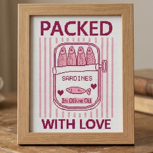 Pu&ograve; includere: Schema a punto croce in una cornice di legno. Il design presenta uno sfondo a righe rosa e bianche con un'illustrazione di una scatoletta di sardine. Le parole "PACKED", "SARDINES", "In Olive Oil" e "WITH LOVE" sono ricamate in rosa.