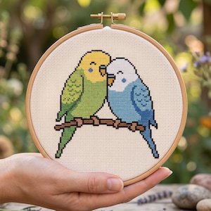 K&ouml;nnte beinhalten: Ein runder Stickrahmen aus Holz mit einem Kreuzstichmuster von zwei Wellensittichen, die auf einem braunen Ast sitzen. Ein Vogel ist gelb und gr&uuml;n, der andere wei&szlig; und blau. Beide V&ouml;gel haben die Augen geschlossen und schauen sich an.