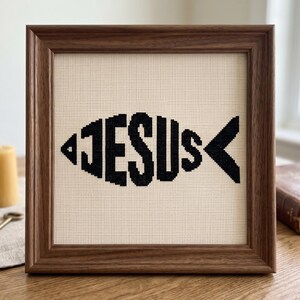 Puede incluir: Un bordado en punto de cruz cuadrado que presenta la palabra JESUS en hilo negro, dispuesta para formar la silueta de un pez. El bordado est&aacute; sobre un fondo de tela de color crema y se encuentra dentro de un marco de madera marr&oacute;n oscuro.