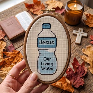 Puede incluir: Un bastidor de bordado ovalado de madera que contiene un dise&ntilde;o de punto de cruz sobre tela color crema. El dise&ntilde;o presenta una botella de agua azul claro con el texto "Jesus" y "Our Living Water" bordado en hilo azul oscuro.