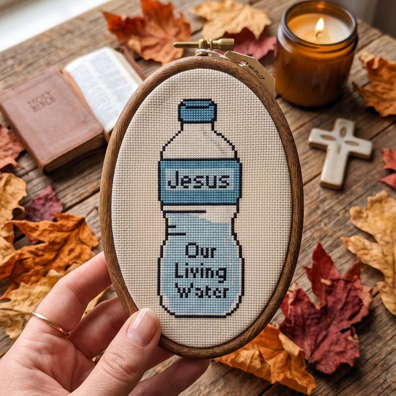 Puede incluir: Un bastidor de bordado ovalado de madera que contiene un dise&ntilde;o de punto de cruz sobre tela color crema. El dise&ntilde;o presenta una botella de agua azul claro con el texto "Jesus" y "Our Living Water" bordado en hilo azul oscuro.
