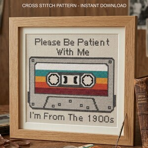 May include: A framed cross stitch pattern featuring a gray cassette tape with horizontal stripes in teal, yellow, orange, and red. The text reads "Please Be Patient With Me" at the top and "I'm From The 1900s" at the bottom in black cross stitch lettering.
