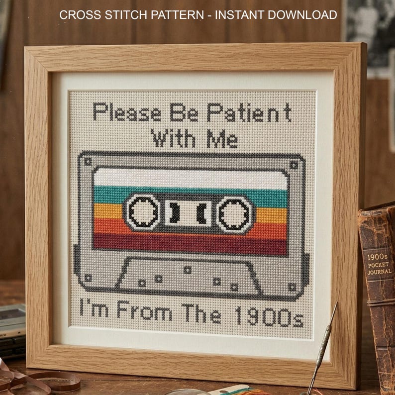 May include: A framed cross stitch pattern featuring a gray cassette tape with horizontal stripes in teal, yellow, orange, and red. The text reads "Please Be Patient With Me" at the top and "I'm From The 1900s" at the bottom in black cross stitch lettering.