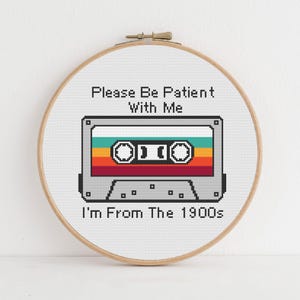 May include: A circular wooden embroidery hoop containing a cross-stitch design of a grey cassette tape with horizontal stripes in teal, yellow, orange, and red. The text reads: Please Be Patient With Me I'm From The 1900s in black pixelated font.