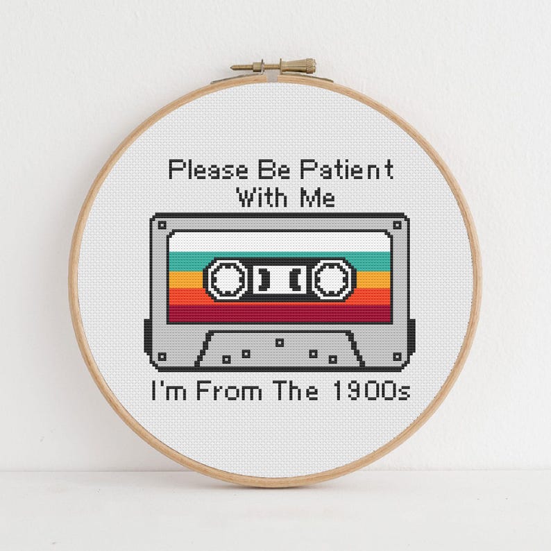 May include: A circular wooden embroidery hoop containing a cross-stitch design of a grey cassette tape with horizontal stripes in teal, yellow, orange, and red. The text reads: Please Be Patient With Me I'm From The 1900s in black pixelated font.