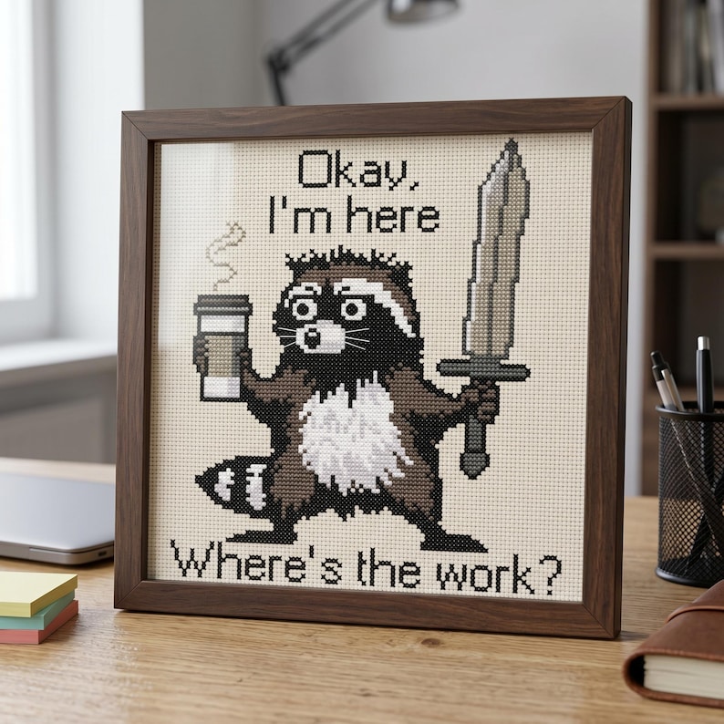 Puede incluir: Imagen de punto de cruz enmarcada en madera oscura. Muestra un mapache sosteniendo una taza de caf&eacute; y una espada. El texto dice: "Okay, I'm here" y "Where's the work?" El mapache es marr&oacute;n, negro y blanco.