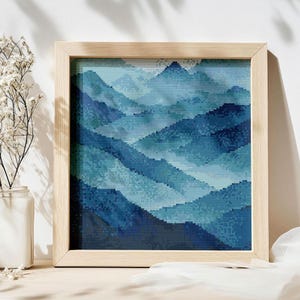 May include: A framed cross-stitch artwork depicting a mountain scene in various shades of blue. The piece is presented in a light wood frame. The mountain range is created using a pixelated technique, giving it a textured look.