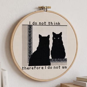 May include: A circular wooden embroidery hoop containing a cross-stitch piece on cream fabric. It depicts two black cat silhouettes facing each other with the text "I do not think" above and "therefore I do not am" below the cats.