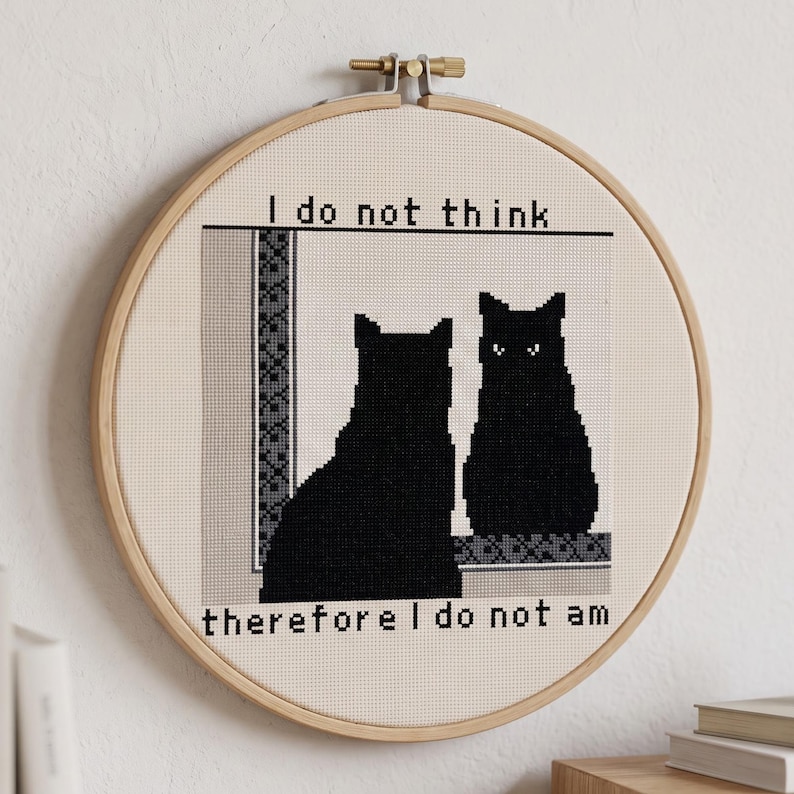 May include: A circular wooden embroidery hoop containing a cross-stitch piece on cream fabric. It depicts two black cat silhouettes facing each other with the text "I do not think" above and "therefore I do not am" below the cats.