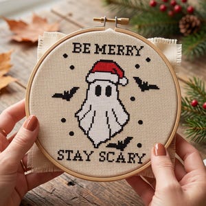 K&ouml;nnte beinhalten: Ein runder Stickrahmen aus Holz mit einem Kreuzstichmotiv auf beigem Stoff. Das Design zeigt ein wei&szlig;es Gespenst mit einer roten Weihnachtsm&uuml;tze, umgeben von schwarzen Flederm&auml;usen und Punkten, mit dem Text BE MERRY und STAY SCARY in Schwarz.