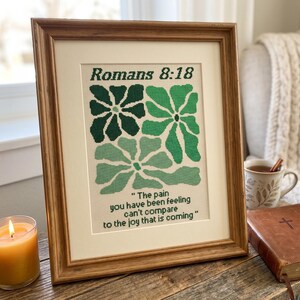 May include: A framed cross-stitch piece with green floral patterns and the text "Romans 8:18". The quote reads, "The pain you have been feeling can't compare to the joy that is coming." The artwork is in a wooden frame, displayed on a wooden surface.