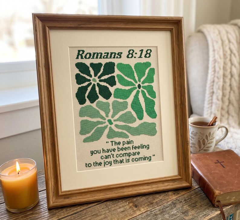 May include: A framed cross-stitch piece with green floral patterns and the text "Romans 8:18". The quote reads, "The pain you have been feeling can't compare to the joy that is coming." The artwork is in a wooden frame, displayed on a wooden surface.