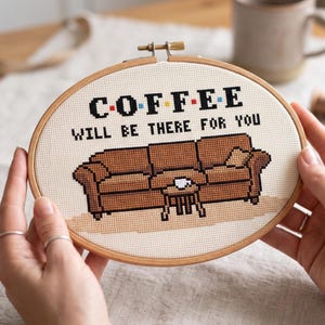 May include: An oval wooden embroidery hoop containing a cross-stitch design on cream fabric. The design features a brown sofa, a small table with a cup, and the black text COFFEE WILL BE THERE FOR YOU with colorful dots between the letters.