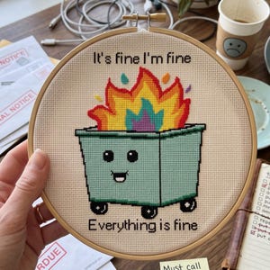 K&ouml;nnte beinhalten: Ein runder h&ouml;lzerner Stickrahmen mit einem Kreuzstichmotiv eines hellgr&uuml;nen M&uuml;llcontainers auf R&auml;dern mit Gesicht, der von gelben, orangefarbenen und violetten Flammen umgeben ist. Der Text lautet It's fine I'm fine und Everything is fine.