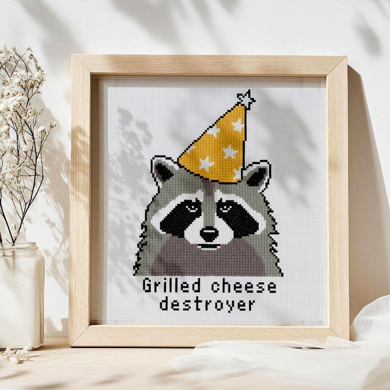 Op de afbeelding: Kruissteekkunst van een wasbeer met een gele feesthoed met witte sterren. De wasbeer heeft een zwart masker en is omlijst in een licht houten frame. De tekst "Grilled cheese destroyer" staat eronder.