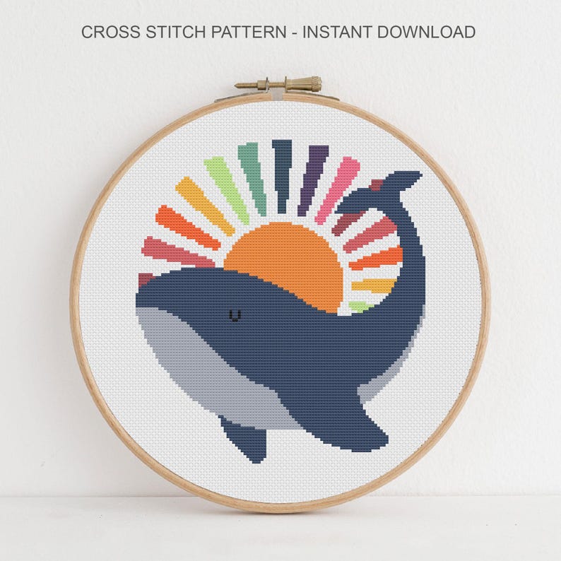 Op de afbeelding: Een kruissteekpatroon met een blauwe walvis met een kleurrijk zonnemotief. De walvis lacht en de zon heeft regenboogkleurige stralen. Het patroon wordt weergegeven in een houten borduurring, met de tekst "CROSS STITCH PATTERN - INSTANT DOWNLOAD" bovenaan.