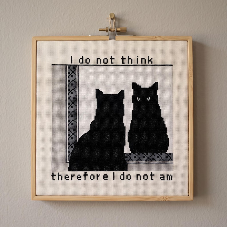 May include: A square cross-stitch embroidery in a light wood frame featuring two black cats. One cat faces away, while the other faces forward with white eyes. The text reads I do not think above and therefore I do not am below the cats.