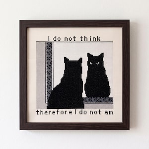 Puede incluir: Un bordado en punto de cruz enmarcado que muestra un gato negro sentado frente a un espejo, con su reflejo visible. El texto "I do not think" está bordado sobre el gato, y "therefore I do not am" está bordado debajo con hilo negro.
